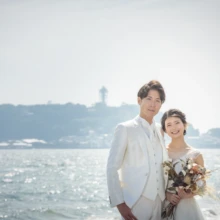 鎌倉 和洋装 K様・K様 2021.05.24 江の島を望む七里ヶ浜で、ブーケを持つウェディングドレスの新婦と白いスーツの新郎が並ぶ鎌倉前撮り