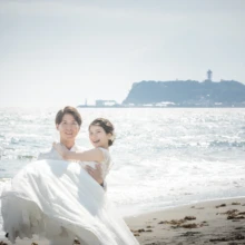 鎌倉 和洋装 K様・K様 2021.05.24 七里ヶ浜で白いスーツ姿の新郎がウェディングドレスの新婦を抱き上げ、江の島を背に笑い合う洋装前撮り