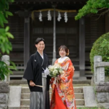 鎌倉 和装 B様・A様 2025.07.12 緑に囲まれた隣の神社の社殿前で、赤い色打掛の新婦と黒紋付袴の新郎がブーケを手に並ぶ