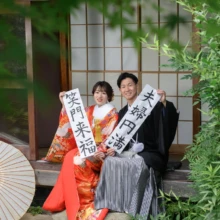 鎌倉 和装 B様・A様 2025.07.12 鎌倉の古民家スタジオの庭で、色打掛の新婦と黒紋付袴の新郎がメッセージ入りのたすきを持って座る