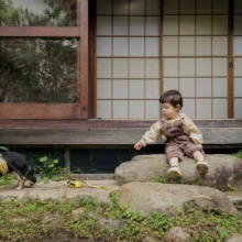 ペット・家族写真 T様 2025.10.24 鎌倉の古民家スタジオの庭で、岩に座る幼い子どもの前を黄色いハーネス姿の黒白の愛犬が歩く自然な家族撮影