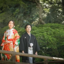 鎌倉 和洋装 H様・A様 2021.03.29 妙本寺の新緑を背景に、白地に赤や緑の文様が入った色打掛姿の新婦と黒紋付袴姿の新郎が木造の欄干前に立つ
