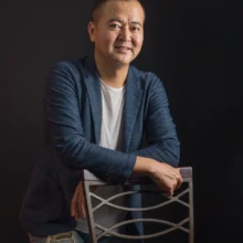 プロフィール写真 I様 2022.09.22 暗いスタジオで、紺色のジャケットとデニム姿の男性が椅子の背に両腕を置き、カメラを見るポートレート