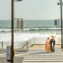 鎌倉和装 H様・H様 2021.03.29 海へ続く横断歩道の中央で、緑と赤の色打掛姿の新婦と黒紋付袴姿の新郎が白い和傘を差して向き合う