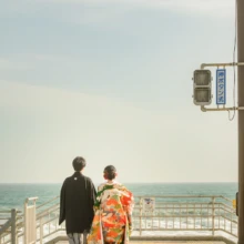 鎌倉和装 H様・H様 2021.03.29 鎌倉高校前の海へ続く階段を、緑と赤の色打掛姿の新婦と黒紋付袴姿の新郎が並んで下りる後ろ姿
