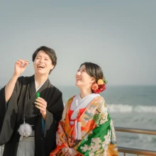 鎌倉和装 H様・H様 2021.03.29 海を背景に、小物を手に掲げて笑う黒紋付袴姿の新郎と、緑と赤の色打掛姿で新郎を見上げる新婦