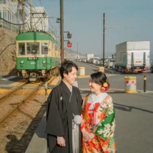 鎌倉和装 H様・H様 2021.03.29 鎌倉高校前の江ノ電線路沿いで、緑と赤の色打掛姿の新婦と黒紋付袴姿の新郎が緑色の車両を背に微笑み合う