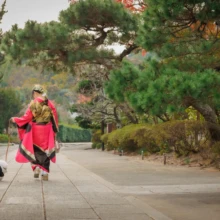 成人式撮影 S様 2022.12.03 秋の建長寺の参道で、赤い振袖姿の女性が両腕を広げ、地面にいる白い愛犬を見守る後ろ姿