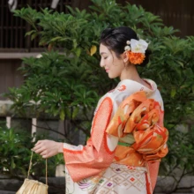 成人式撮影 M様 2023.08.09 鎌倉の神社境内で、橙色の花飾りと華やかな帯結びを見せ、金色の和装バッグを持つ成人式の女性