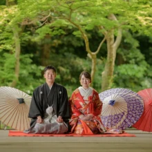 鎌倉 和装 M様・H様 2021.06.02 妙本寺の本堂前に正座し、白・紫・赤の和傘を背に微笑む朱赤の色打掛の新婦と黒紋付袴の新郎