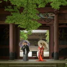 鎌倉 和装 M様・H様 2021.06.02 妙本寺の二天門で紫と白の和傘を差し、向かい合って立つ朱赤の色打掛の新婦と黒紋付袴の新郎