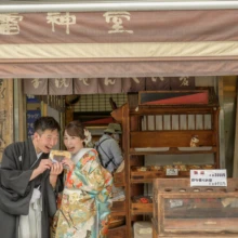 鎌倉 和装 M様・H様 2021.06.02 鎌倉の昔ながらの店先で、朱赤の色打掛の新婦と黒紋付袴の新郎が顔を寄せて笑う街歩き前撮り