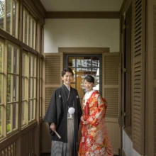 横浜 和装 Y様・O様 2022.04.14 木製の窓が連なる山手西洋館の廊下で、黒紋付袴の新郎と赤い色打掛の新婦が並ぶ横浜和装前撮り