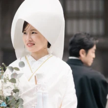 建長寺で綿帽子と白無垢をまとい、ブーケを手に微笑む花嫁の一枚|縁グラフィの姉妹ブランドLuca 建長寺でブーケを手に微笑む綿帽子と白無垢姿の花嫁と、背景に佇む黒紋付姿の新郎