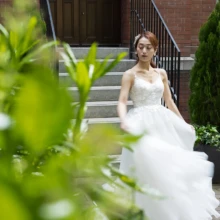 赤レンガのクラシカルな建築と鮮やかな緑に、純白のウェディングドレスが映える一枚。ドレスの動きを活かし、丸の内らしい上品で華やかな雰囲気に|縁グラフィの姉妹ブランドLuca 東京・丸の内の赤レンガ建築を背景に、緑越しにウェディングドレス姿の花嫁を撮影した前撮り写真