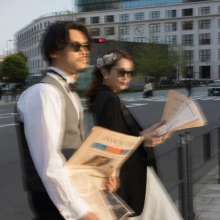スローシャッターによる動きのブレを活かした、印象的な一枚。サングラスと新聞を合わせ、丸の内の都会的なロケーションをファッション誌のような雰囲気で切り取ったウェディングフォト|縁グラフィの姉妹ブランドLuca 東京・丸の内の街並みを背景に、サングラスと新聞を手にした新郎新婦をスローシャッターで撮影したウェディングフォト