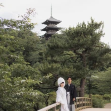 三溪園を象徴する三重塔を望む橋の上で。歴史ある景観と豊かな緑を背景に残す、白無垢と黒紋付の和装フォトウェディング|縁グラフィの姉妹ブランドLuca 横浜・三溪園の三重塔と緑豊かな日本庭園を背景に、白無垢姿の花嫁と黒紋付姿の新郎が橋に佇む和装ウェディングフォト