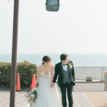 由比ヶ浜の海を望む街並みで、手をつないで歩くふたりの自然なウェディングフォト|縁グラフィの姉妹ブランドLuca 由比ヶ浜の海を背景に、手をつないで横断歩道を渡るウェディングドレス姿の花嫁とタキシード姿の新郎