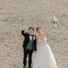 由比ヶ浜の砂浜で弾ける笑顔を残した、ふたりらしい自然体のウェディングフォト|縁グラフィの姉妹ブランドLuca 由比ヶ浜の砂浜で手をつなぎ、笑顔でブーケを掲げるウェディングドレス姿の花嫁とタキシード姿の新郎