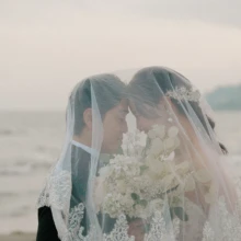 由比ヶ浜の海辺でベールに包まれながら寄り添う、ふたりのロマンチックなウェディングフォト|縁グラフィの姉妹ブランドLuca 由比ヶ浜の海を背景に、レースのベールに包まれて額を寄せ合う新郎新婦のウェディングフォト