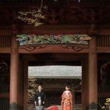 鎌倉和洋装S様・S様 2019.12.23 歴史ある寺院の大きな山門を背景に、黒紋付袴の新郎と赤い色打掛の新婦が並ぶ鎌倉前撮り