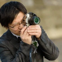 鎌倉和洋装S様・S様 2019.12.23 海辺の洋装前撮りで、眼鏡を掛けた新郎が小型カメラのファインダーをのぞき撮影する様子