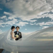 鎌倉和洋装S様・S様 2019.12.23 青空と海を背景に、長いウェディングベールに包まれて寄り添うタキシードの新郎とドレス姿の新婦
