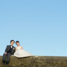 鎌倉和洋装S様・S様 2019.12.23 澄んだ青空の下、海沿いの堤防に並んで腰掛けるタキシードの新郎とウェディングドレスの新婦