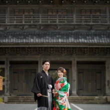 鎌倉 和装 T様・U様 2020.09.29 光明寺の荘厳な山門を背景に、緑と赤の色打掛姿の新婦と黒紋付袴の新郎が並ぶ鎌倉前撮り
