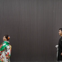 鎌倉 和装 T様・U様 2020.09.29 鎌倉の黒い縦格子壁を背景に、色打掛の新婦と黒紋付袴の新郎が距離を取って歩く横長構図