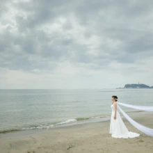 鎌倉 和洋装 T様・Y様 2021.03.20 七里ガ浜の海と江の島を背景に、ロングベールを海風になびかせて砂浜に立つウェディングドレス姿の新婦