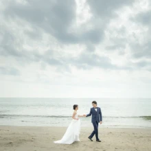 鎌倉 和洋装 T様・Y様 2021.03.20 曇り空の七里ガ浜で、ウェディングドレス姿の新婦と青いタキシード姿の新郎が手をつないで砂浜を歩く