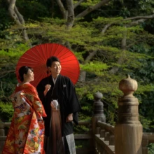 鎌倉 和洋装 T様・Y様 2021.03.20 妙本寺の新緑を背景に、赤い和傘のそばで笑い合う赤い色打掛姿の新婦と黒紋付袴姿の新郎