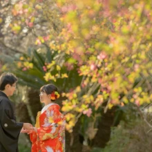 鎌倉 和洋装 T様・Y様 2021.03.20 妙本寺の春の花を背景に、赤い色打掛姿の新婦と黒紋付袴姿の新郎が向き合い、手を取り合う