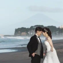鎌倉和洋装 Y様・Y様 2026.08.08 七里ガ浜の朝の海辺で、江の島と富士山を背景に額を寄せるタキシードの新郎とウェディングドレスの新婦