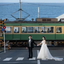 鎌倉和洋装 Y様・Y様 2026.08.08 海を望む七里ヶ浜の踏切で江ノ電を背景に手をつなぐ、ウェディングドレス姿の花嫁とタキシード姿の新郎