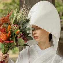 鎌倉 和装 N様N様 2020.11.02 妙本寺の柱のそばで、綿帽子と白無垢姿の新婦が赤やオレンジの花を束ねたブーケを見つめる花嫁ソロ