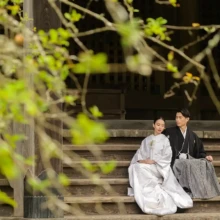 鎌倉 和装 N様N様 2020.11.02 妙本寺の石段に腰掛け、白無垢姿の新婦が黒紋付袴姿の新郎に寄り添う様子を新緑の枝越しに捉えた鎌倉前撮り