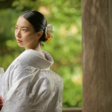 鎌倉 和装 N様N様 2020.11.02 妙本寺の新緑を背景に、白無垢姿の新婦が赤やオレンジの花を束ねたブーケを持ち、髪飾りを見せて振り返る