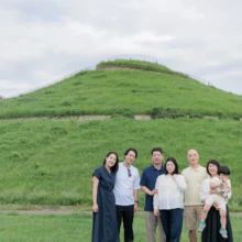家族写真 M様 2025.09.21 緑の丘が広がる公園で、乳幼児を含む三世代の家族が肩を寄せて並ぶ秋のファミリーロケーション撮影