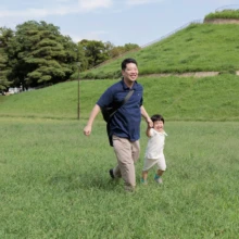家族写真 M様 2025.09.21 緑の丘を背景に、広い芝生を笑顔で駆ける父親と、その後ろを追いかける幼い子どもの親子撮影