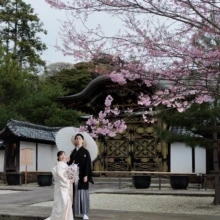 鎌倉 和装 S様・B様 2026.04.01 建長寺の唐門と濃い桃色の桜を背景に、石段に立つ白無垢の新婦と白い番傘を差す黒紋付袴の新郎