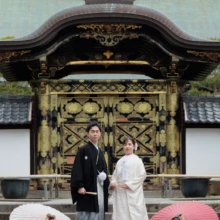 鎌倉 和装 S様・B様 2026.04.01 建長寺の装飾豊かな唐門前で、白と赤の番傘を両脇に置き並んで立つ白無垢の新婦と黒紋付袴の新郎