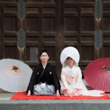 鎌倉 和装 S様・B様 2026.04.01 建長寺の木造建築前で、白と赤の番傘を両脇に置き、綿帽子姿の新婦と黒紋付袴の新郎が正座する姿