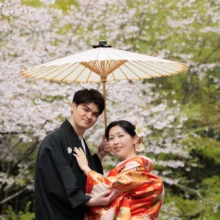 鎌倉 和装 M様・I様 2026.04.02 淡い桜を背景に白い番傘の下で、新婦の肩を抱いて寄り添う黒紋付袴の新郎と赤い色打掛の新婦