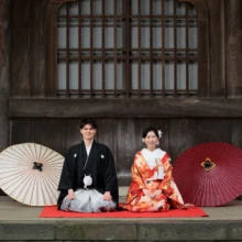 鎌倉 和装 M様・I様 2026.04.02 建長寺の木造建築前で、白と赤の番傘を両脇に置き、赤い敷物の上に正座する和装の新郎新婦