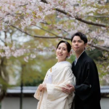 鎌倉 和装 T様・K様 2026.04.05 円覚寺の満開の桜を背景に、白無垢の新婦を黒紋付袴の新郎が後ろから支えて寄り添う上半身のカット