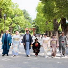 七五三撮影 I様 2026.06.01 新緑に包まれた鶴岡八幡宮の参道を、七五三の女の子と和装姿の家族7人が横一列で歩く様子