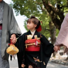 七五三撮影 I様 2026.06.01 黒い着物姿の女の子が両側の家族と手をつなぎ、黄色い巾着を持って参道を歩く七五三の一場面