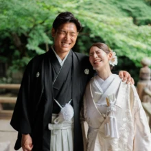 鎌倉 和装 I様・Y様 2026.07.05 緑豊かな妙本寺の境内で、新郎が白無垢の新婦の肩を抱き、二人で顔を寄せて笑う自然な瞬間